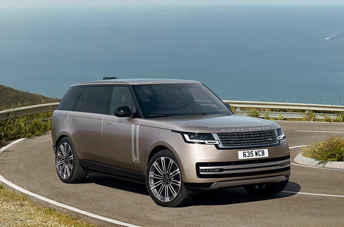 A champagne gold Range Rover driving along a winding coastal road with hills and the sea in the background.