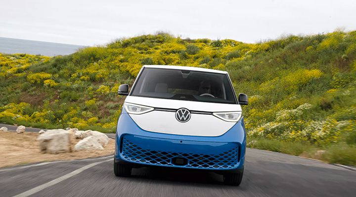 Front view of a two-tone blue and white Volkswagen ID. Buzz electric van driving on a road next to a hill covered in yellow flowers.