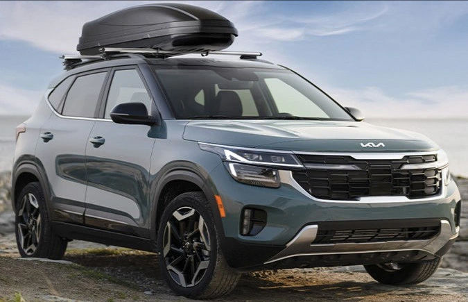  A sage green Kia Seltos SUV with a black cargo box on its roof rack, parked on a sandy surface near the water.