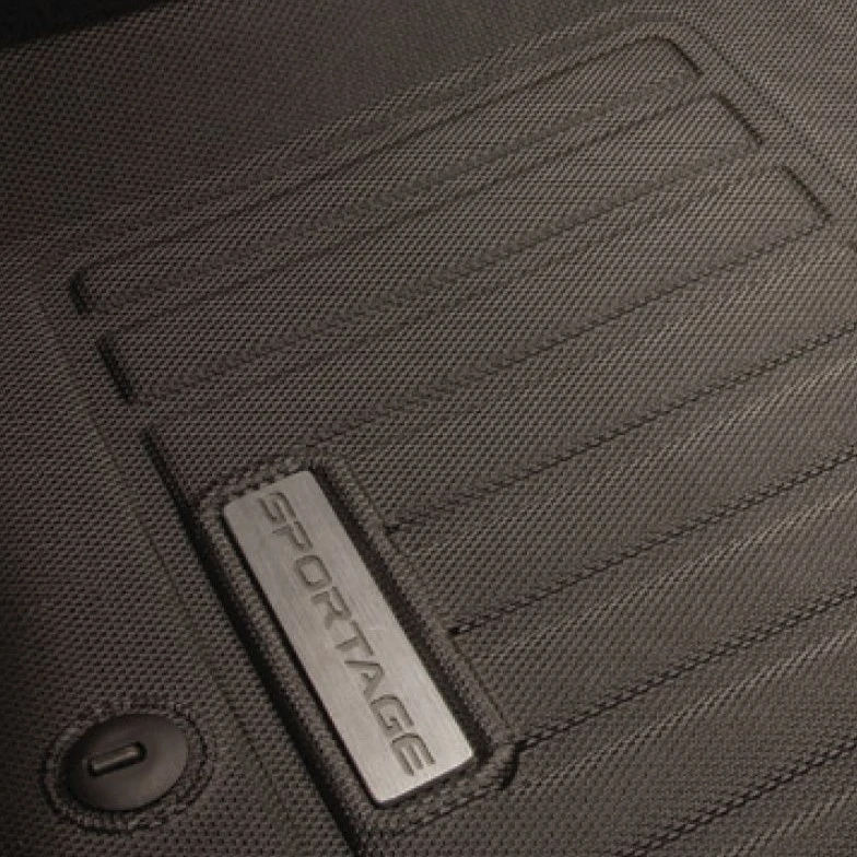 A close-up of a black, textured all-weather floor mat featuring a silver plaque with the word "SPORTAGE" on it.