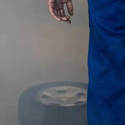 A close-up on the dirty hand of a person in blue work coveralls, with a tire visible through a hazy or smoky background.