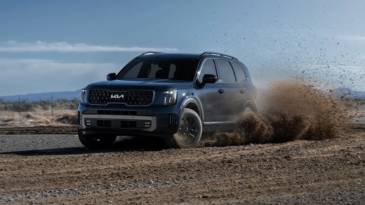A dark blue Kia Telluride SUV turning on a dirt road, kicking up a large cloud of dust and gravel.