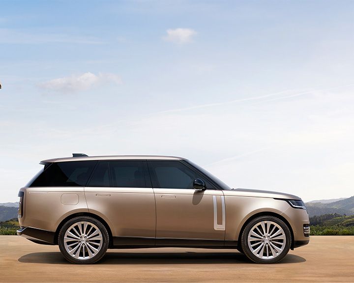 Side profile of a champagne gold Range Rover against a light blue sky with wispy clouds and distant hills.