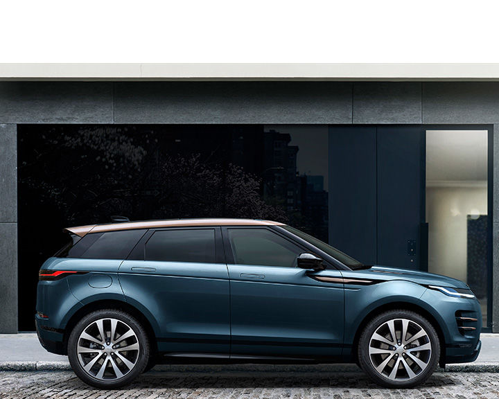 Side profile of a metallic teal Land Rover Evoque with a contrasting copper-colored roof, parked in front of a modern dark building facade.