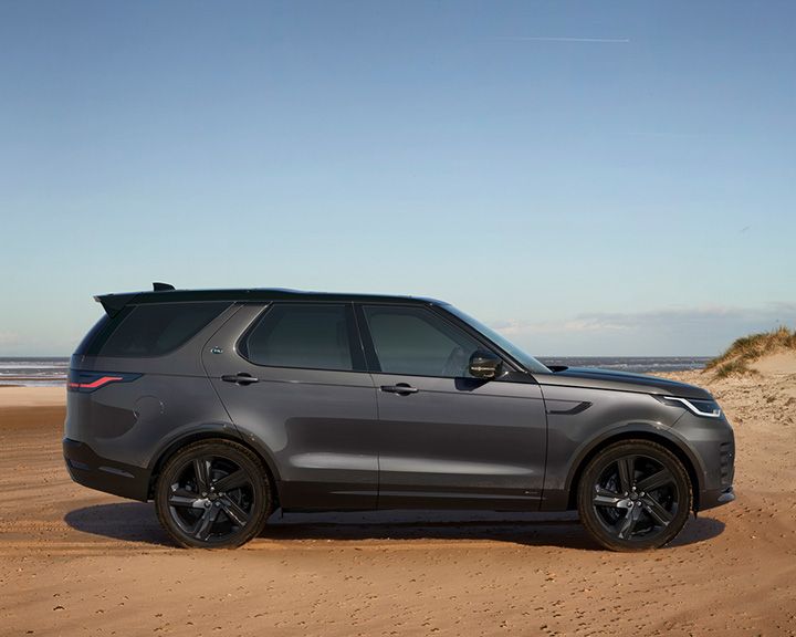 Side profile of a dark grey Land Rover Discovery parked on a sandy beach with the ocean and a dune in the background under a clear sky.