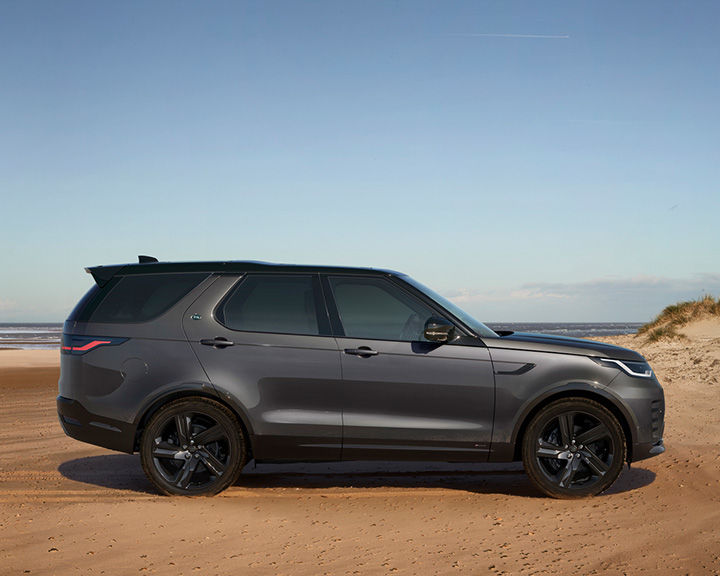 Side profile of a dark grey Land Rover Discovery parked on a sandy beach with the ocean and a dune in the background under a clear sky.