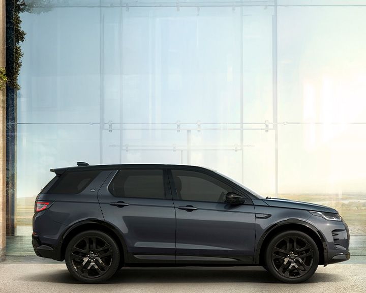 Side profile of a dark grey Land Rover Discovery Sport with a black roof, parked in front of a large glass window reflecting a bright outdoor scene.