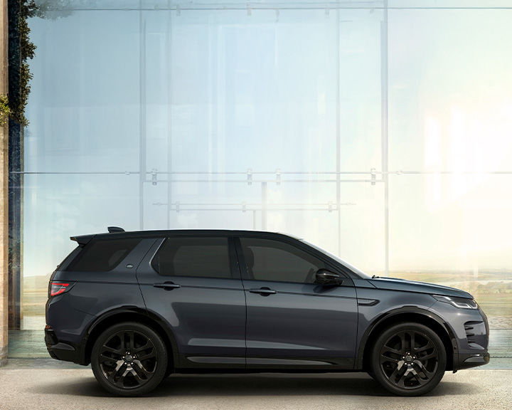 Side profile of a dark grey Land Rover Discovery Sport with a black roof, parked in front of a large glass window reflecting a bright outdoor scene.