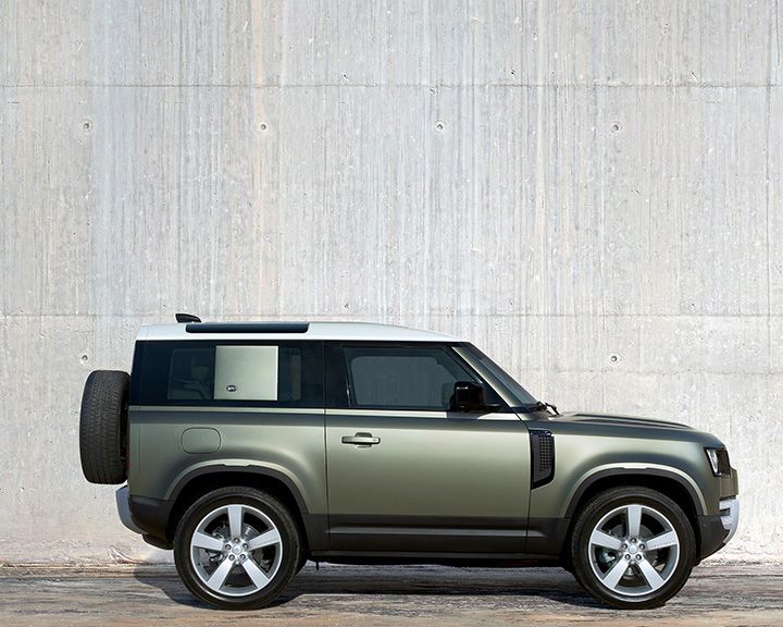 Side profile of a metallic olive green Land Rover Defender 90 with a white roof and a spare tire mounted on the rear, parked against a plain concrete wall.