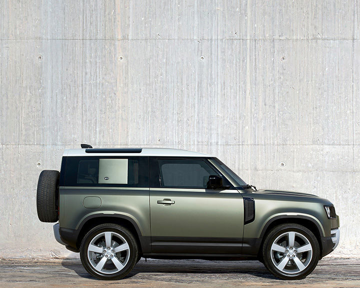 Side profile of a metallic olive green Land Rover Defender 90 with a white roof and a spare tire mounted on the rear, parked against a plain concrete wall.