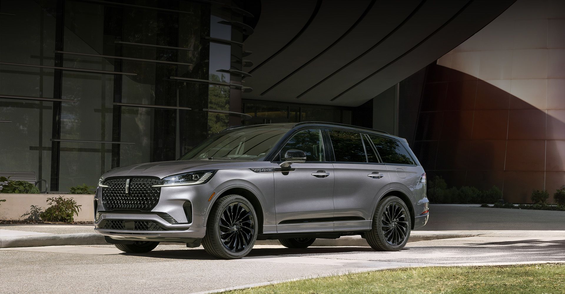 A silver-grey SUV with black wheels is parked on an asphalt driveway in front of a modern building with large glass windows and an overhanging architectural feature. The car is angled slightly towards the left, showcasing its sleek design.