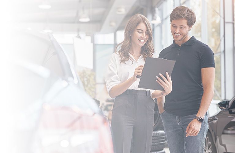 A smiling female salesperson and a male customer look at a clipboard together inside a bright car dealership showroom.