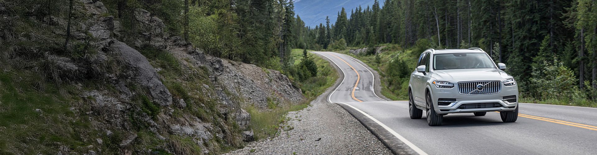 A light-colored Volvo SUV drives along a winding road, flanked by coniferous trees and mountains in the background under a cloudy sky.