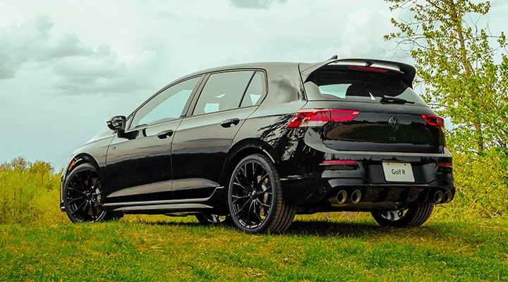 Rear view of a black Volkswagen Golf R with spoiler and quad exhaust, parked on a grassy field.