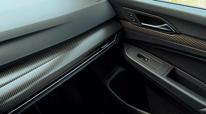 Close-up of a car's interior trim, showing a carbon fiber pattern with gold accents and door controls.