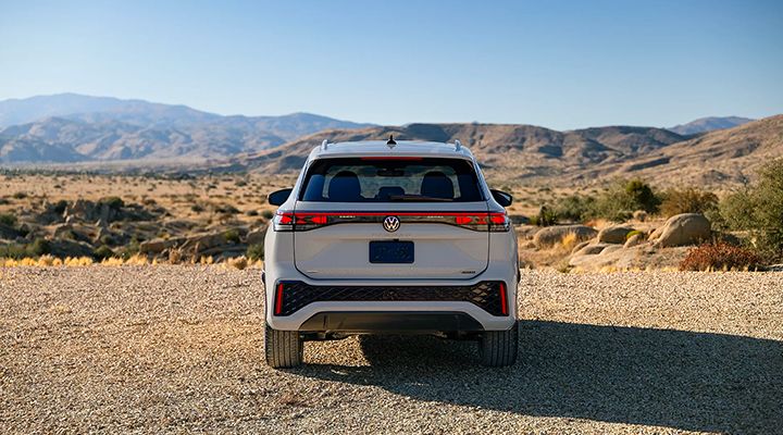Rear view of a light grey Volkswagen SUV parked on a rocky ground, with a desert landscape and mountains in the background.