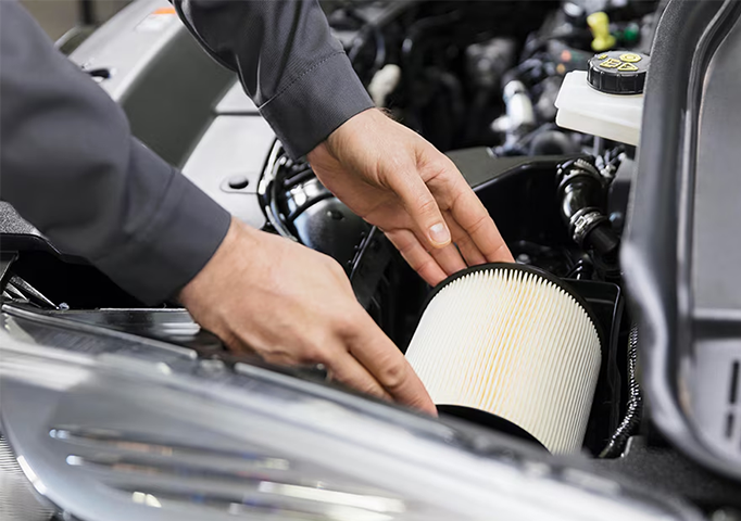 Close-up of a mechanic wearing a grey uniform replacing a round white air filter in a car's engine bay. The mechanic's hands are carefully placing the new filter into the housing.