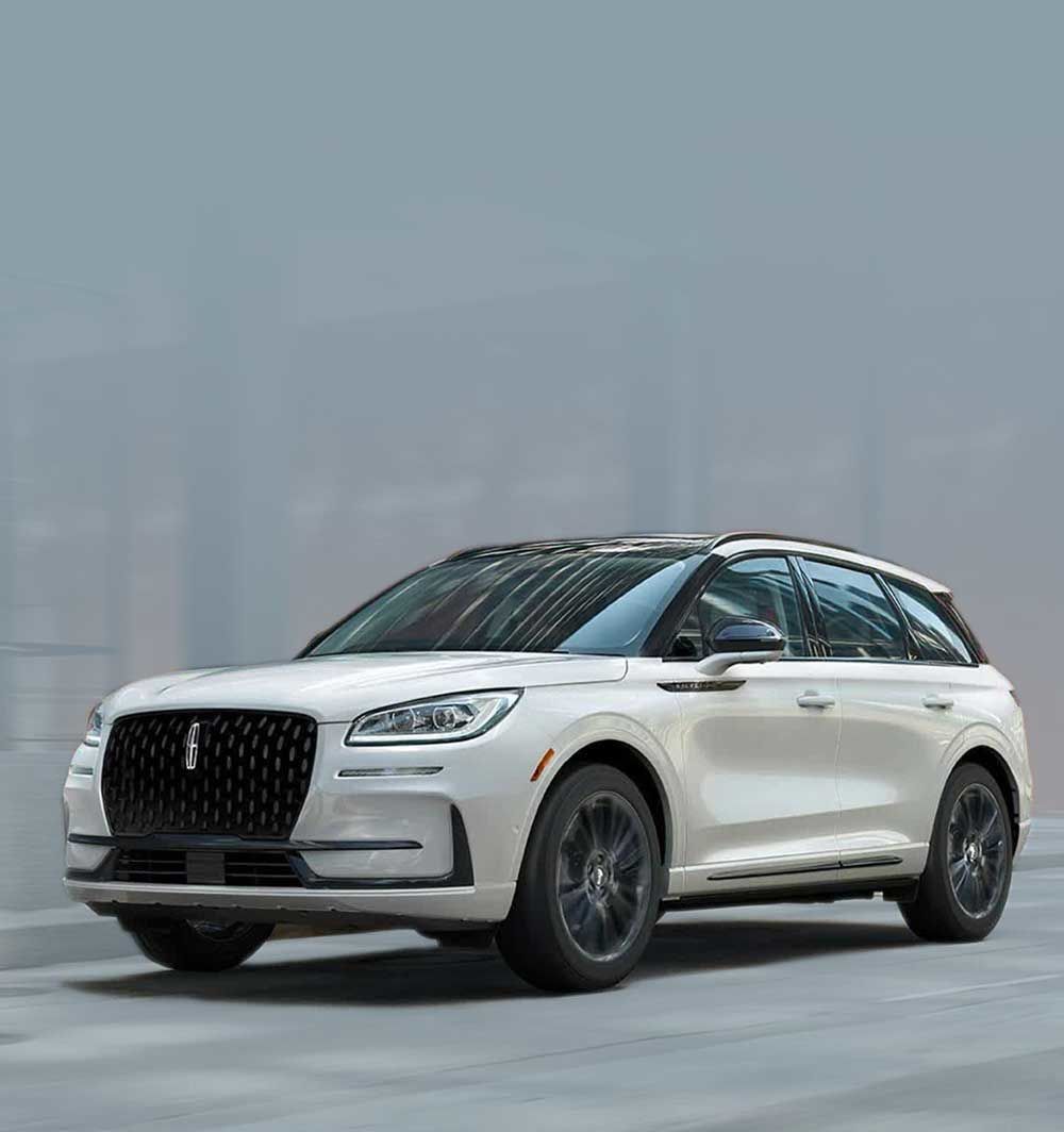  A white Lincoln Corsair SUV drives on a city street, with a red brick building and modern structures in the background. The daytime running lights are on.