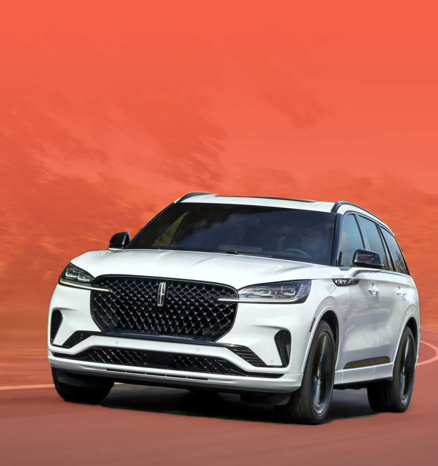 A white Lincoln Aviator SUV drives on a winding road lined with trees. The car is seen from the front, with motion blur indicating movement.