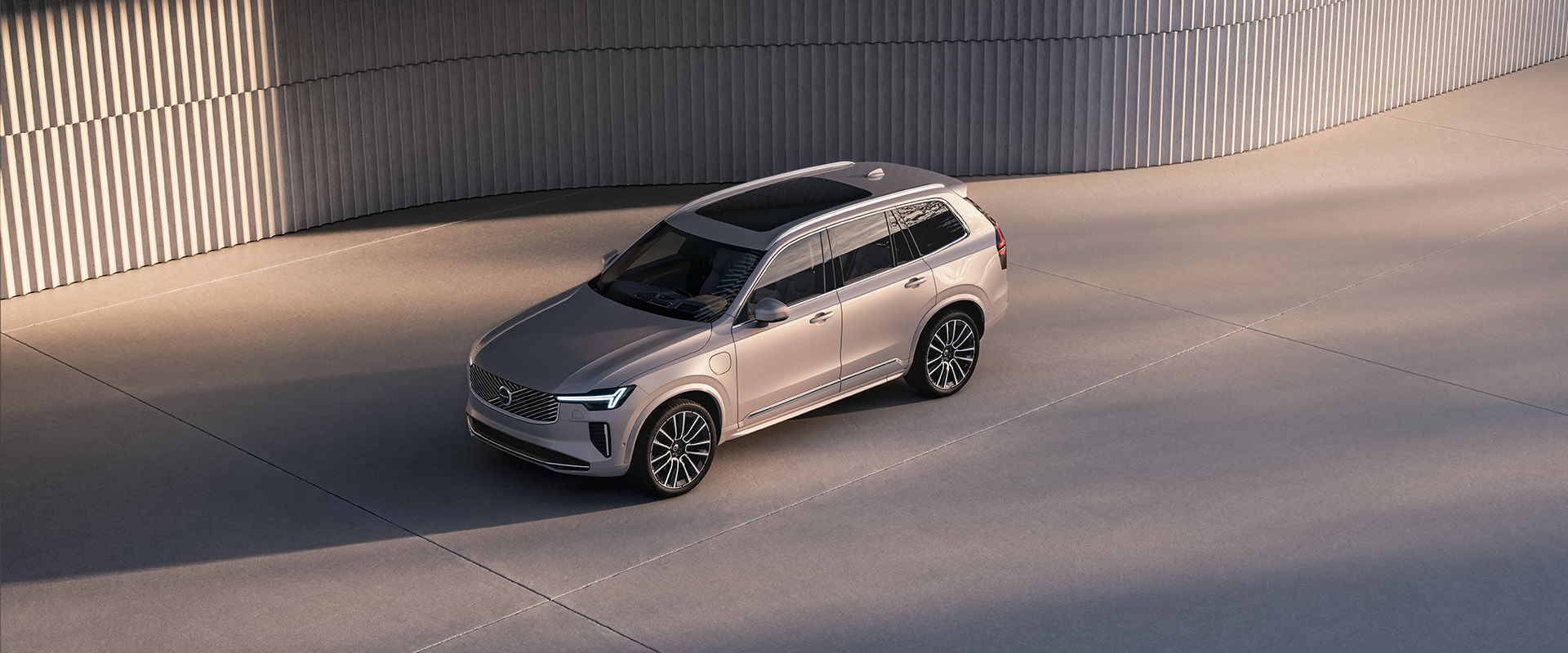 A high-angle view of a silver Volvo EX90 parked on a concrete surface next to a modern, ribbed wall.