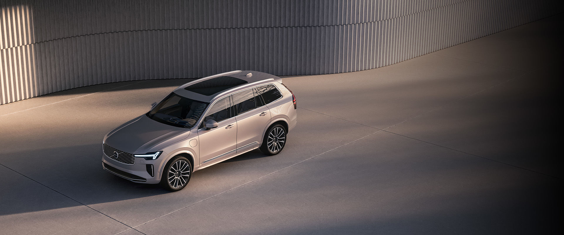 A high-angle view of a silver Volvo EX90 parked on a concrete surface next to a modern, ribbed wall.