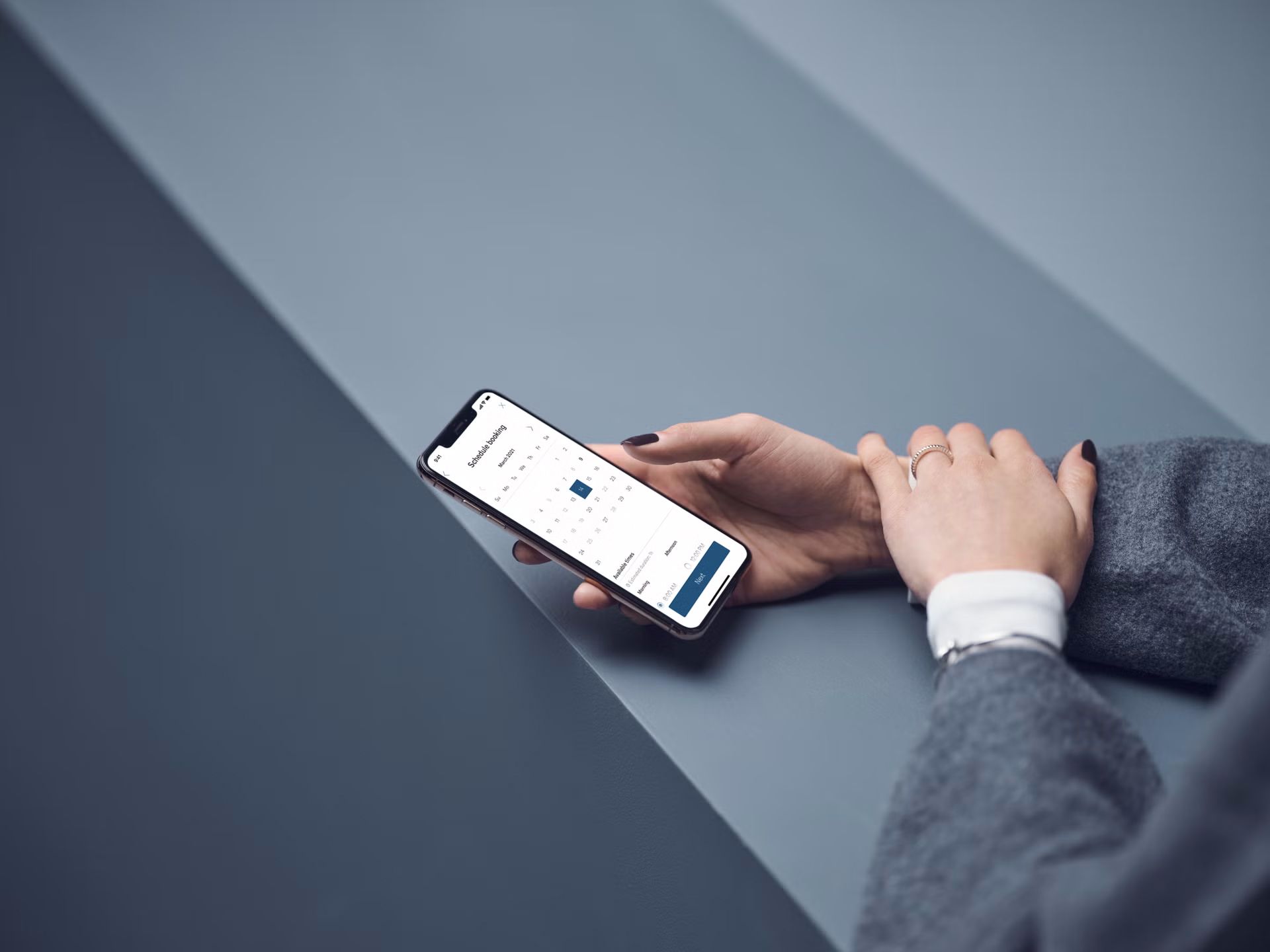 A person in a grey blazer holds a smartphone, viewing a calendar to book a service appointment.