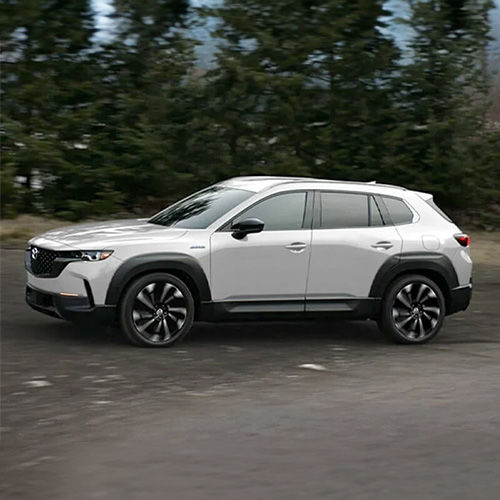 A white CX-50 Hybrid drives alongside trees
