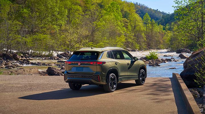 Rear three-quarter view of an olive green Volkswagen SUV parked near a river, with lush greenery in the background.
