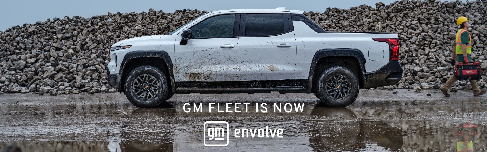 A side-profile photograph of a mud-splattered white Chevrolet Silverado EV parked at a rugged industrial site with a large pile of rocks in the background. A worker in a yellow hard hat and safety vest walks past the truck carrying a red Milwaukee tool case. Large white text centered at the bottom reads "GM FLEET IS NOW" above the GM Envolve logo.