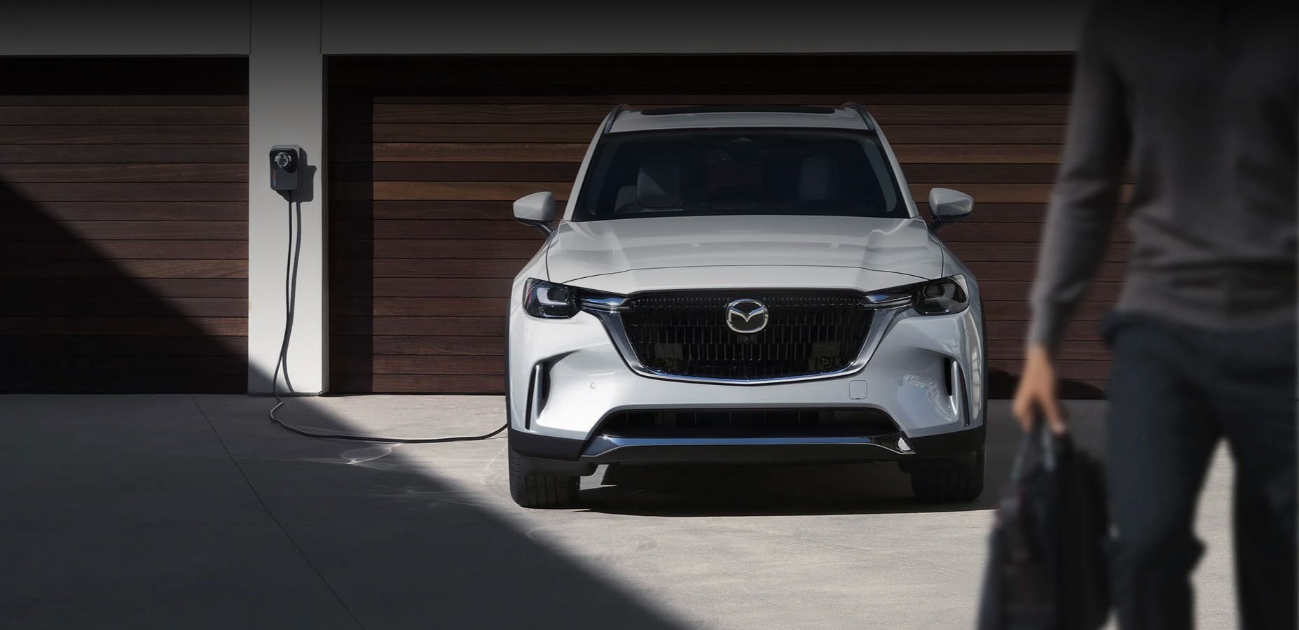Person with tote bag walks away from CX-90 PHEV, plugged in and charging infront of two–door garage.
