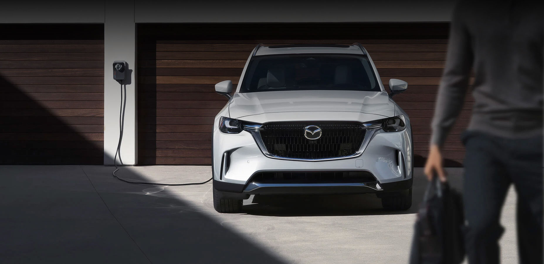 Person with tote bag walks away from CX-90 PHEV, plugged in and charging infront of two–door garage.