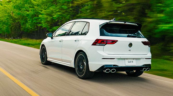Dynamic rear view of a white Volkswagen Golf R driving on a country road, showing the roof spoiler and quad exhaust.