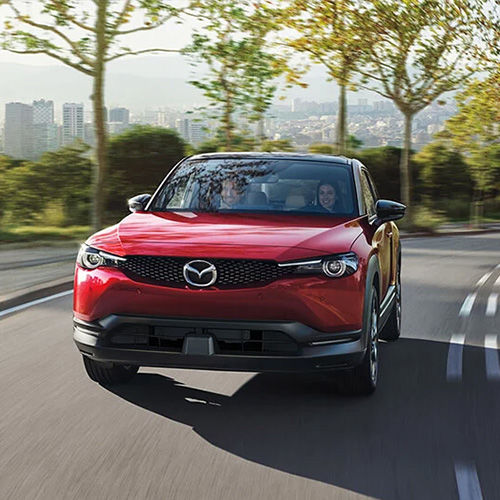 MX-30 EV driving up an escapment road lined by trees that overlooks a city. 