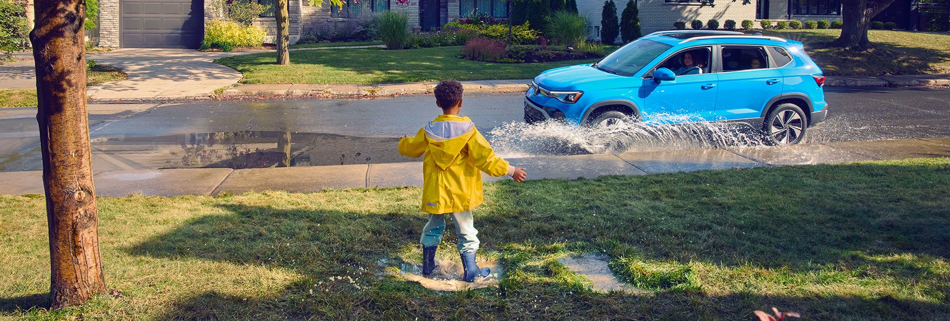 A child in a yellow raincoat splashes in a puddle on a grassy lawn as a bright blue SUV drives through a large puddle on a suburban street.