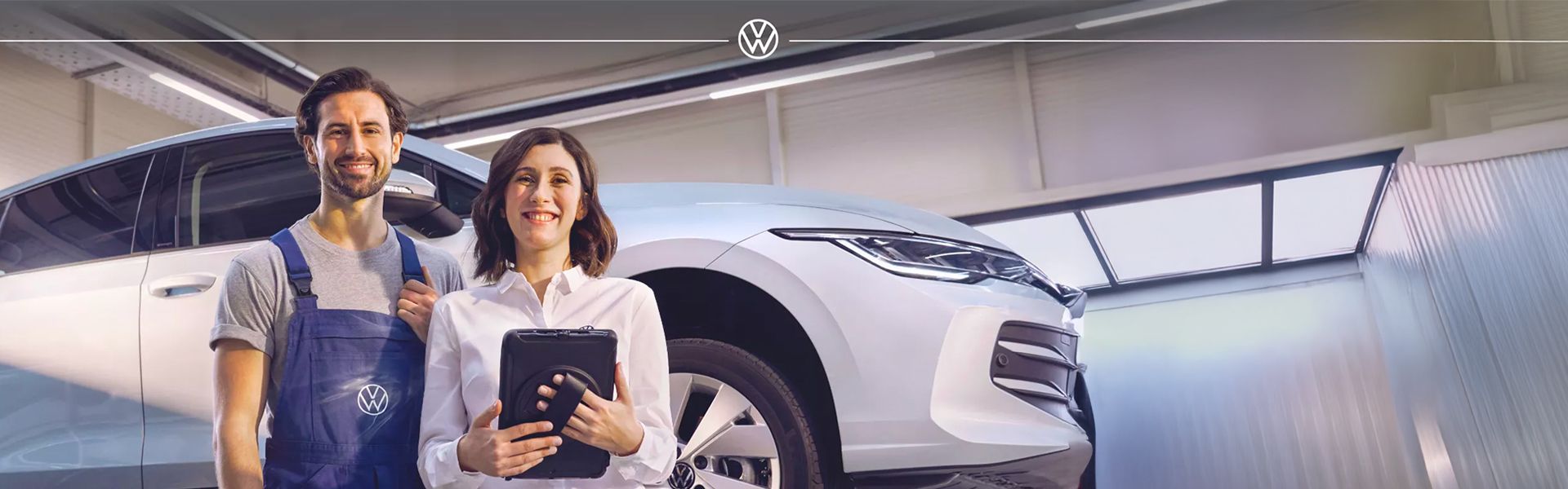 A smiling female service advisor holding a tablet and a male technician in blue overalls stand in front of a white Volkswagen in a bright service bay.