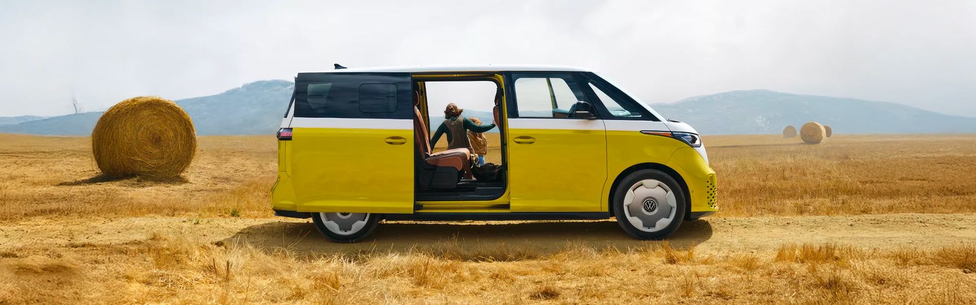 A yellow and white Volkswagen ID. Buzz with its side door open, parked in a rural field filled with large hay bales under a cloudy sky.