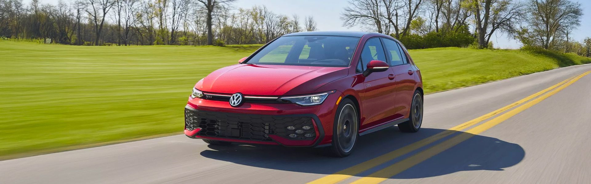 Une voiture à hayon Volkswagen Golf GTI rouge roule rapidement sur une route de campagne asphaltée, avec un arrière-plan de champs verts.