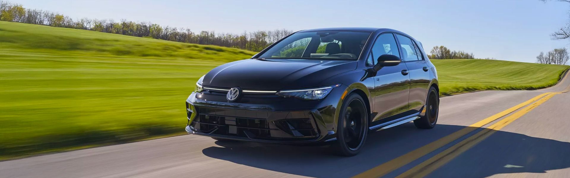 A black Volkswagen Golf R hatchback drives on an asphalt country road, flanked by rolling green hills.
