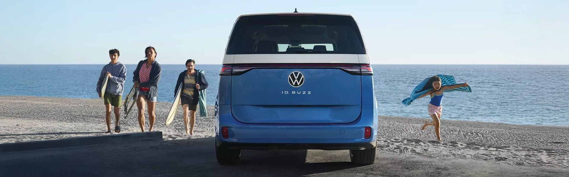 Rear view of a blue and white Volkswagen ID. Buzz electric van parked on a beach. Three people with surfboards walk away from it, and a child runs to the right.