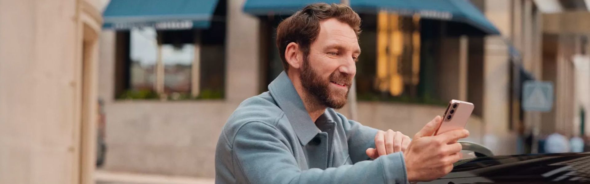 A smiling man leaning on the roof of a black car looks at and uses his pink smartphone in an urban setting.