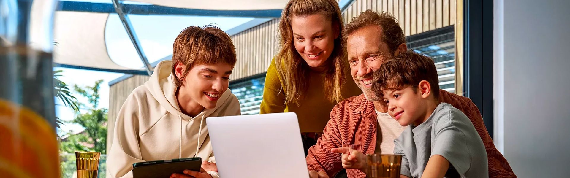A smiling family—mother, father, and two children (a teenager and a young boy)—looking together at a laptop. The setting is bright, modern, and friendly, suggesting a family activity in an indoor/outdoor space.