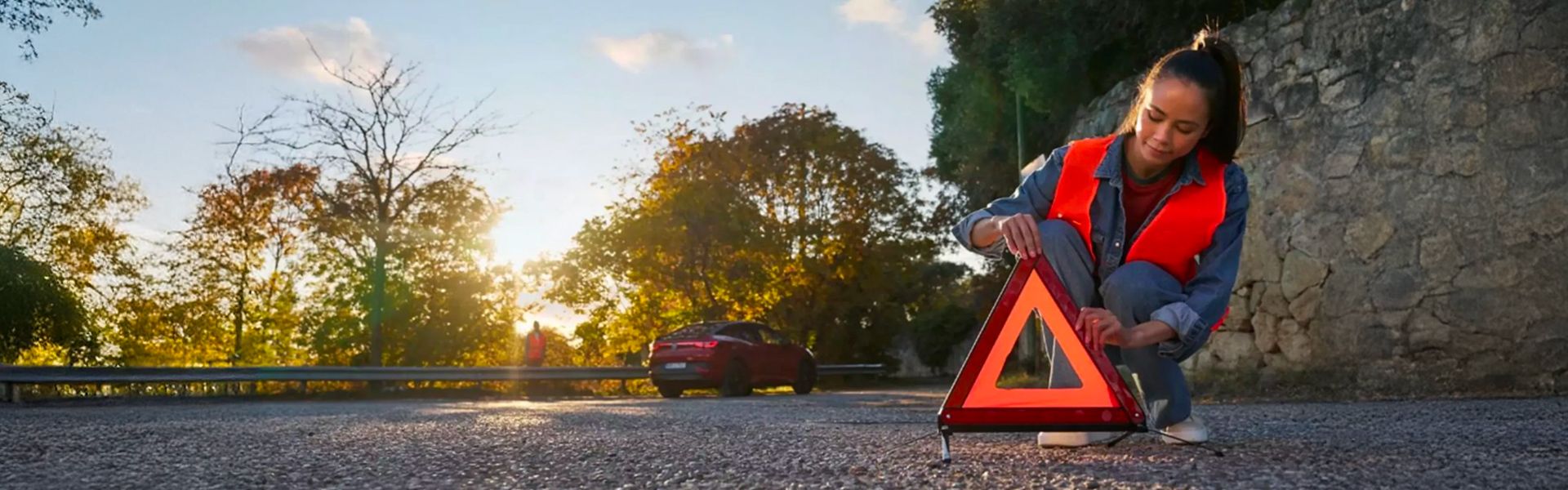 A woman wearing an orange safety vest sets up a warning triangle on the side of a road at sunset. A red car is visible in the background.