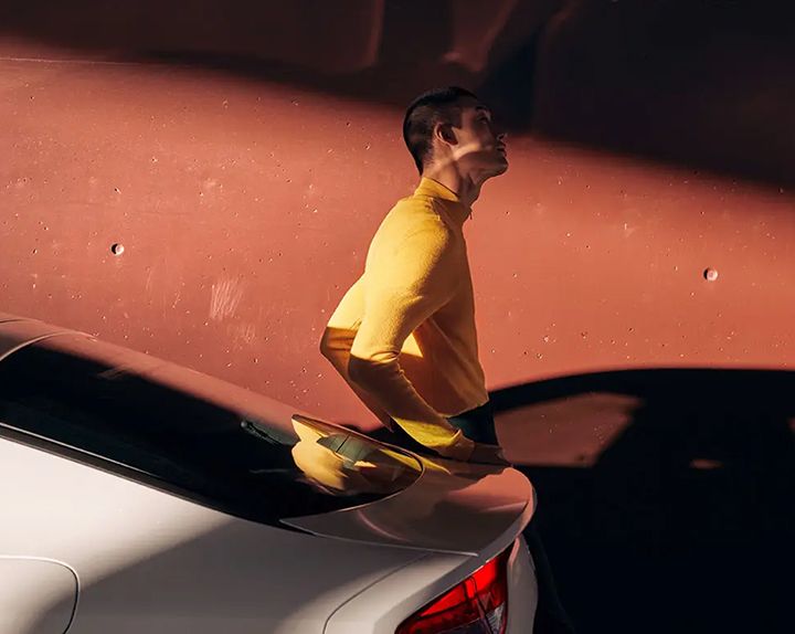 A person in a yellow sweater is leaning on the trunk of a white car, looking up.