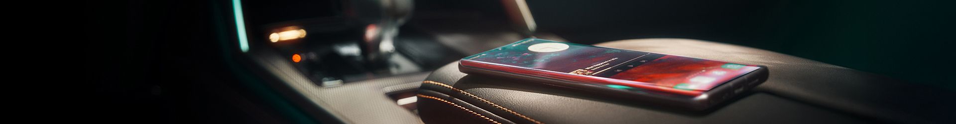A close-up shot of the interior of a Mazda car. A smartphone rests on the leather-stitched armrest, with the dashboard and gear shifter visible in the background, illuminated by a blue ambient light.