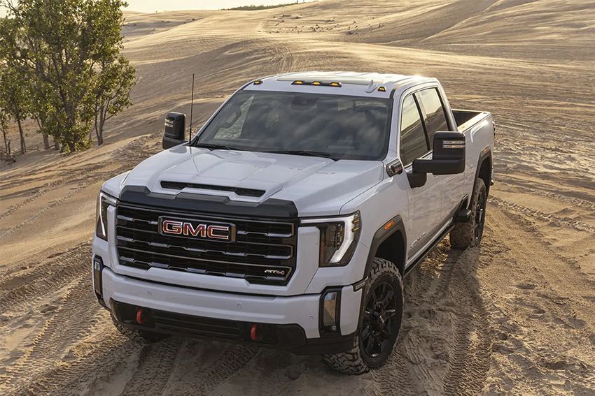 High-angle shot of a white GMC Sierra Heavy-Duty (HD) crew cab truck, likely an AT4X trim, driving or parked in a bright, sandy desert or dune environment.
