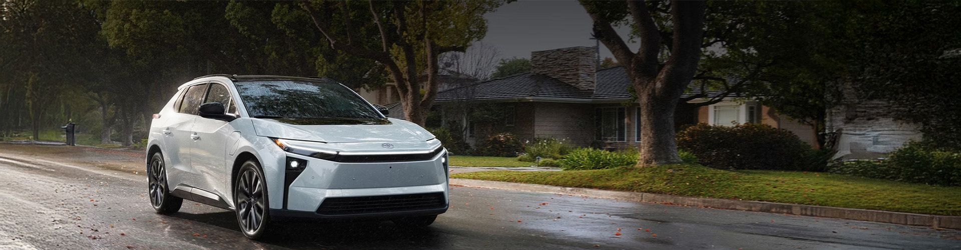 A white Toyota bZ4X electric SUV driving on a wet residential street lined with trees.