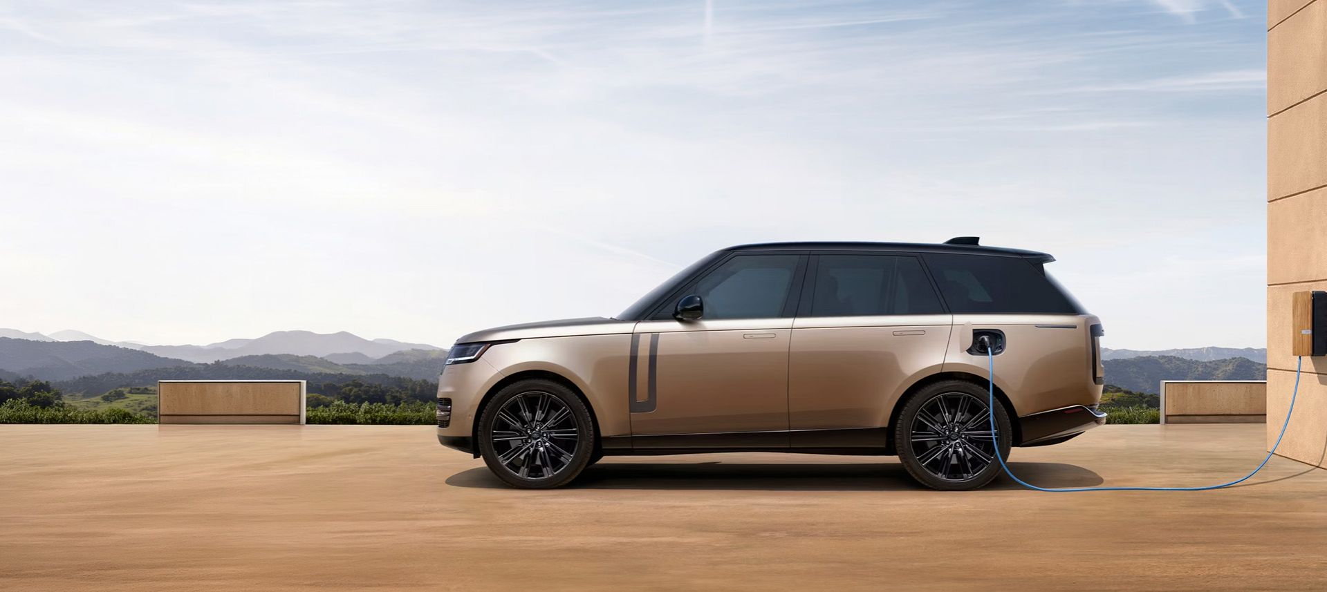 Side profile of a bronze Range Rover charging at an electric station facing the mountains.