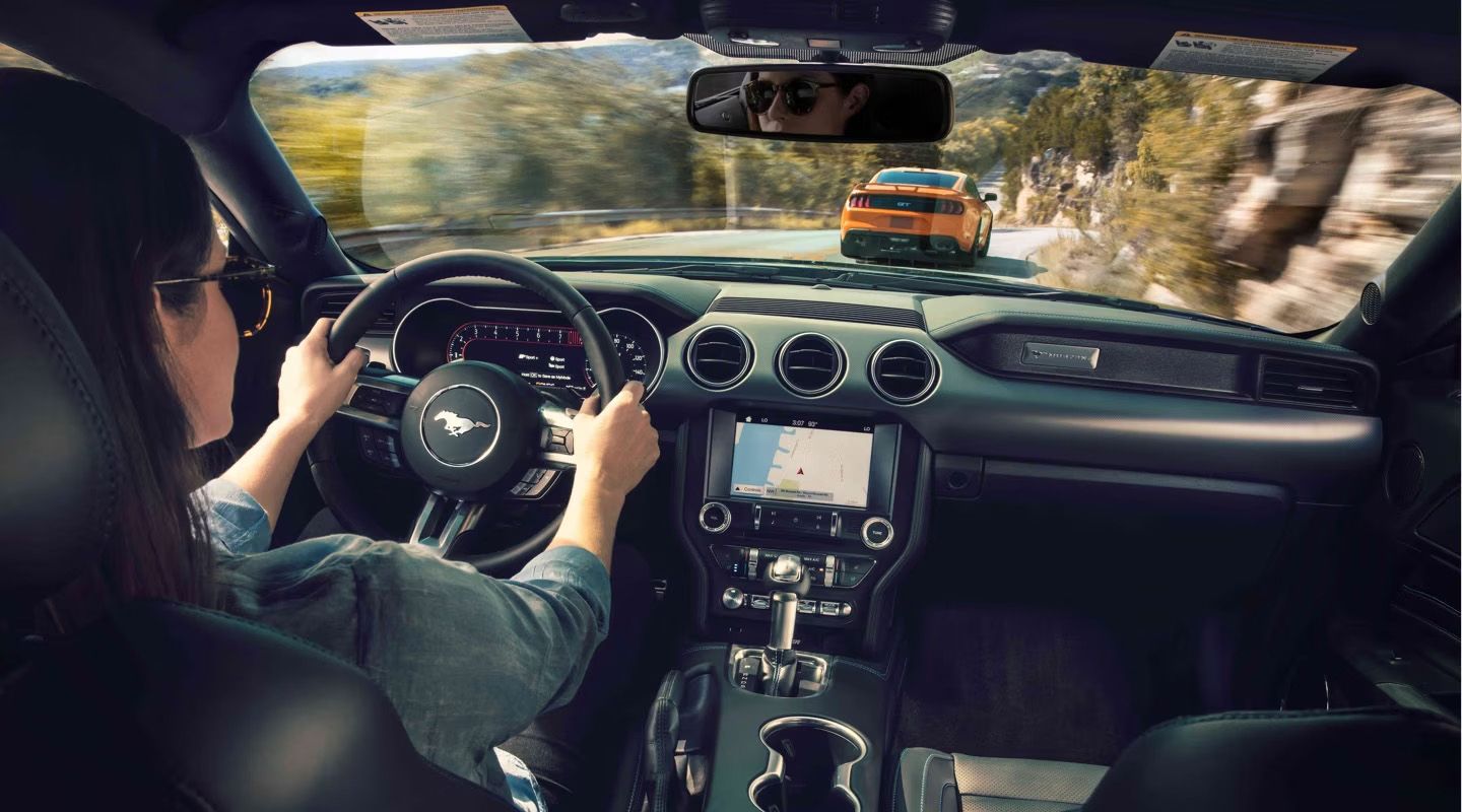 A person is driving a modern car, likely a Ford Mustang, with their hands on the steering wheel. Another orange sports car is visible in the rearview mirror, and the car is on a winding road with trees.