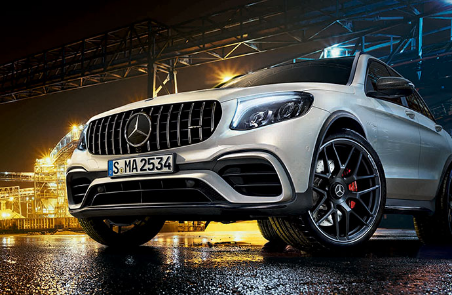 Un SUV Mercedes-AMG blanc stationné de nuit devant un site industriel illuminé.
