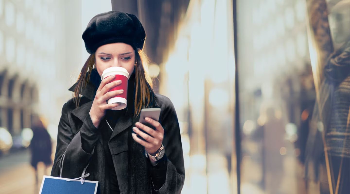 A woman wearing a black coat and a black beret stands outdoors, looking at her phone while drinking from a red and white cup. A blurred storefront is in the background.